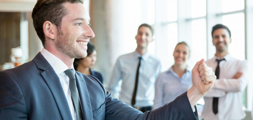 Cheerful leader motivating his business team. Handsome young politician telling his plan and showing fist as symbol of power. Positive business people standing in background. Strong company concept