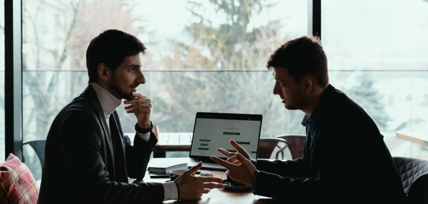 Two businessmen talking about new opportunities sitting with laptop at desk, planning project, considering business offer, sharing ideas while drinking coffee together