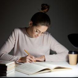 Busy freelancer rewrites infrormation into notepad, prepares article for publication, reads books, writes some notes in organizer, drinks takeaway coffee, sits in darkness, wears round big spectacles