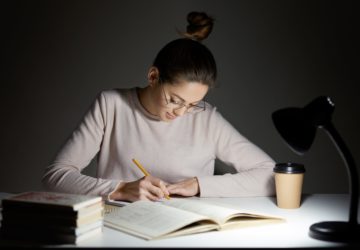 Busy freelancer rewrites infrormation into notepad, prepares article for publication, reads books, writes some notes in organizer, drinks takeaway coffee, sits in darkness, wears round big spectacles