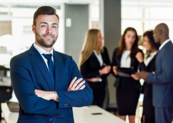 Caucasian businessman leader looking at camera in modern office with multi-ethnic businesspeople working at the background. Teamwork concept. Young man with beard wearing blue suit.