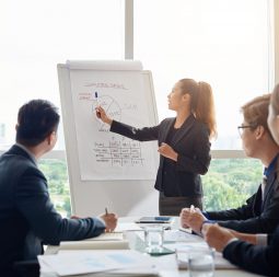 Attractive Asian businesswoman with ponytail pointing at diagram on marker board while holding working meeting in spacious boardroom