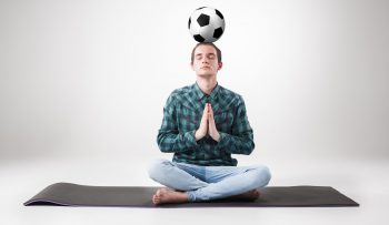 Portrait  of young man, practicing yoga with football ball on gray background. concept of continuous soccer practice